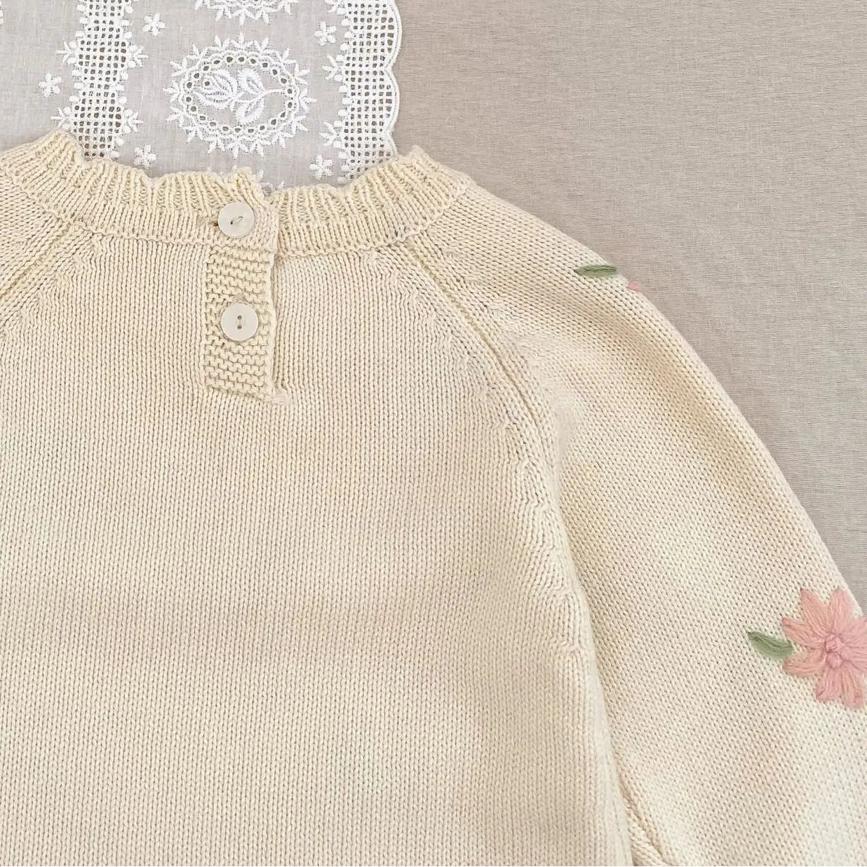 Beige baby romper with floral embroidery on a textured surface with lace and flowers.