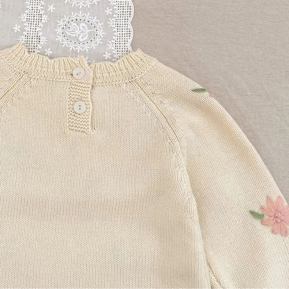 Beige baby romper with floral embroidery on a textured surface with lace and flowers.